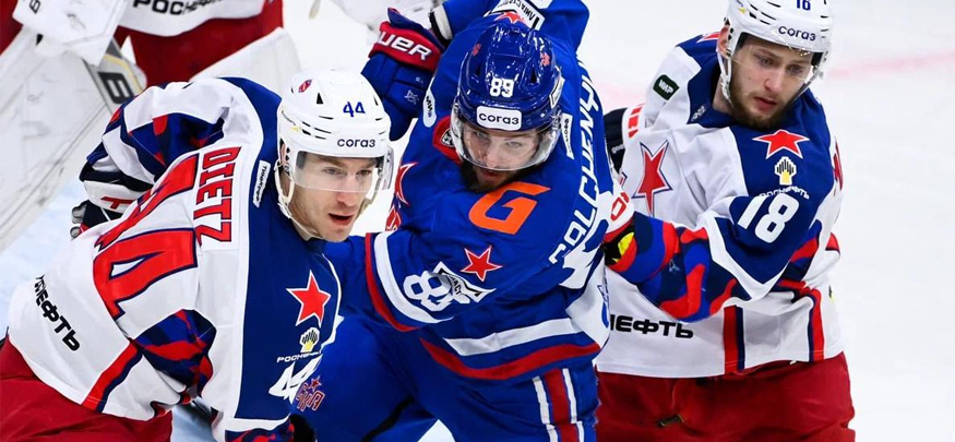 На изображении три хоккеиста в моменте борьбы за шайбу на льду. Один из них в синей форме, двое в белой. Все игроки носят шлемы и оснащены хоккейной экипировкой