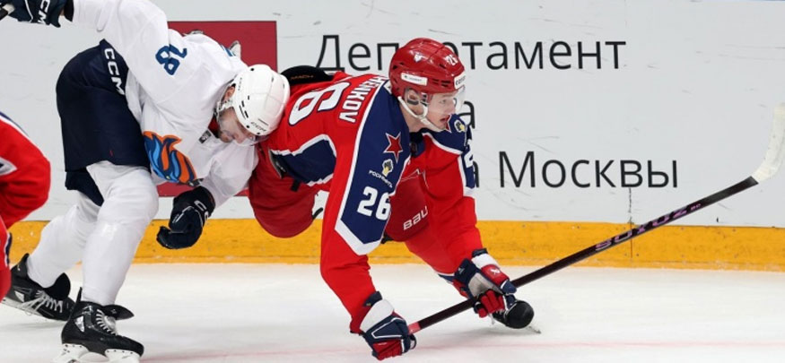 Хоккейный матч: игрок в красной форме, наклонившись, ведет шайбу, пытаясь избежать соперника в бело-синей форме. Они борются за контроль вблизи борта с надписью о московском департаменте спорта