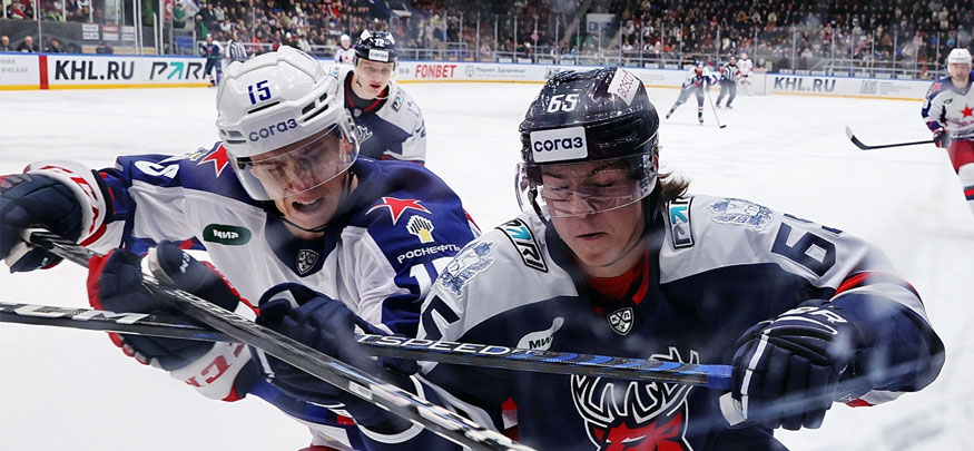 На изображении два хоккеиста в динамичной борьбе за шайбу на льду. Они одеты в разные хоккейные формы, один — в темную, другой — в светлую форму. На заднем плане видны трибуны с болельщиками и реклама