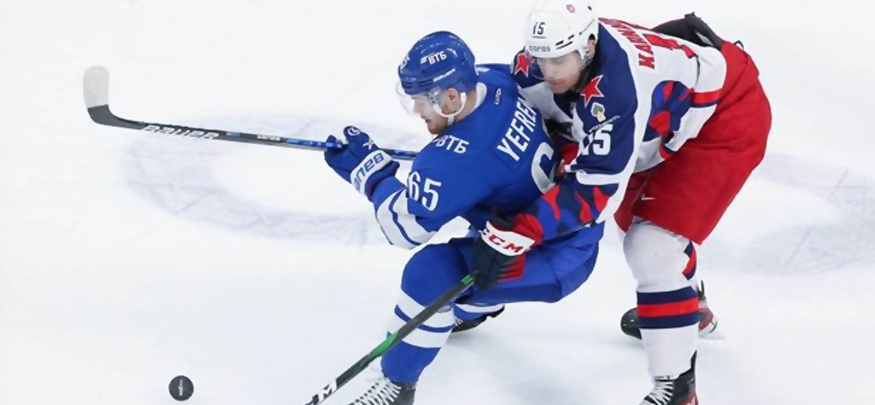 На изображении два хоккеиста в момент борьбы за шайбу на льду. Игрок в синей форме с клюшкой в руках защитно удерживается от соперника в бело-красной форме, при этом враг пытается отобрать шайбу